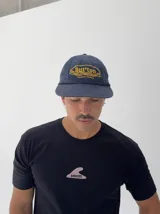 Gorro tipo gorra de béisbol color azul marino, con visera corta y diseño bordado en amarillo con la palabra "But'ton" y la frase "Cruise to Victory".