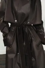 Gabardina larga de efecto encerado en color marrón oscuro, con cuello solapa y cierre frontal cruzado con botones. Presenta mangas largas con puños vueltos en pana, bolsillos laterales y cinturón ajustable de pana. Incluye pinza en la espalda.