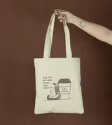 Bolso de mano tipo tote bag color crudo con asas largas y estampado con texto y dibujo de una persona recostada sobre una taza de café.