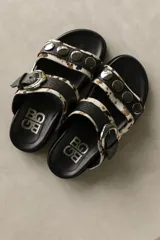 Sandalias de cuero de doble tira, con estampa animal print tipo vaca en tonos blanco, negro y marrón, adornadas con tachas metálicas en la tira superior y una hebilla grande en la tira inferior.