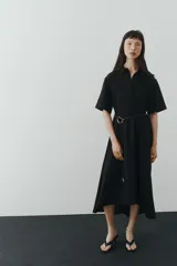 Vestido midi negro con cuello camisero, mangas cortas y botones ocultos. Incluye un cinturón fino con hebilla metálica dorada en la cintura y falda evasé de corte midi.