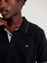 Chomba de manga corta color negro con cuello y tapeta interior en color gris claro. Presenta un pequeño bordado en el pecho y detalles de ribete en gris en el cuello y las mangas.