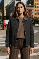 Campera negra de diseño capitoneado con cuello de solapa en contraste color marrón. Presenta cierre frontal con cremallera y botones a presión, mangas largas con puños ajustables y un corte recto.