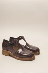 Zapato tipo guillermina de cuero charol color marrón oscuro, con tira T y hebilla ajustable en el lateral. Posee puntera cuadrada y suela de goma color caramelo con taco bajo.