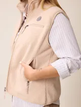Chaleco de sherpa color beige con cierre frontal completo, cuello alto y bolsillo con cierre en el pecho que incluye un detalle circular con logo.
