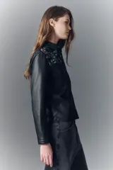 Camisa entallada de símil cuero negro con cuello camisero, botones a presión con logo y bordados florales con lentejuelas en la parte superior delantera.