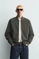 Cazadora bomber de tejido técnico ligero color marrón oscuro, con corte regular fit, cuello, puños y ruedo de rib elástico, bolsillos frontales de vivo y cierre frontal con cremallera.
