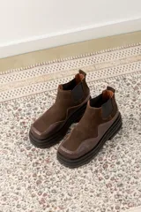 Botas tipo chelsea de caña baja, confeccionadas en una combinación de cuero gamuzado y cuero texturizado en tono marrón oscuro. Presentan una suela track de plataforma gruesa y elásticos laterales negros.
