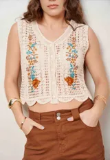 Chaleco de crochet color beige, sin mangas, con escote en V y botones frontales. Cuenta con bordados florales en tono marrón, celeste y ocre en la parte delantera y diseño con ondas en el ruedo.