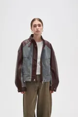 Campera estilo bomber con diseño color block, combinando paneles de denim lavado con mangas y detalles en cuero sintético color marrón oscuro. Presenta cuello alto con tira ajustable, cierre frontal con botones a presión y bolsillos frontales tipo parche.