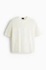 Remera de malla color blanco hueso, de manga corta, con cuello redondo y hombros caídos. Presenta un diseño de tejido perforado con un número grande grabado en el frente.