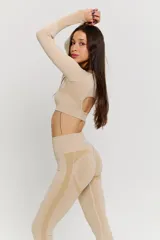 Conjunto deportivo de dos piezas en color beige, compuesto por un top de manga larga con aberturas para pulgares y calzas de tiro alto. Ambas prendas presentan un diseño sin costuras con detalles texturizados y el logo de la marca estampado en el frente del top.