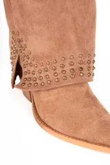 Bota de caña alta estilo slouchy confeccionada en gamuza sintética de color negro, con punta fina y taco bajo. Presenta solapa superior plegada con apliques de tachas metálicas plateadas y una tira con hebilla decorativa en la parte superior de la caña.