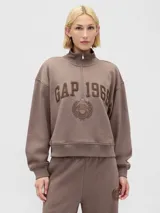 Buzo deportivo de algodón color marrón topo, con cuello alto y medio cierre frontal. Presenta el logo de la marca y el año 1969 bordados en el frente, junto con un emblema circular con un corazón. Posee mangas largas con puños elásticos y un corte holgado.