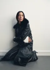 Abrigo largo de estilo trench confeccionado en cuero sintético negro, con diseño recto, cuello con solapa y detalle de cuello de pelo sintético extraíble. Cuenta con cierre frontal de botones, cinturón ajustable con hebilla, bolsillos laterales y mangas largas con trabillas.