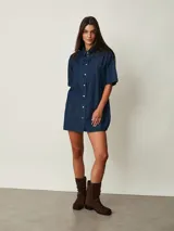 Vestido corto de denim azul oscuro, con cuello camisero, mangas cortas, cierre frontal de botones y dos bolsillos en el pecho.
