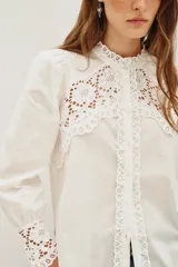 Camisa negra de algodón con detalles de encaje calado en el pecho, hombros y puños. Presenta cuello alto con terminación de encaje, cierre frontal con botones y mangas largas abullonadas.