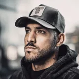 Gorra tipo trucker de color negro, confeccionada en cuero sintético en la parte frontal y visera, con paneles traseros de malla. Presenta un parche rectangular en el centro con un diseño gráfico bordado en color negro sobre fondo beige.