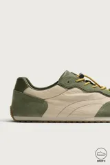 Zapatilla deportiva barefoot fabricada en combinación de piel y tejido, en tonos verde oliva y beige, con cordones elásticos amarillos y suela de goma flexible.
