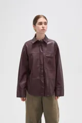 Sobrecamisa de cuero sintético en tono bordó oscuro, con cuello clásico, cierre frontal oculto y un bolsillo aplicado en el pecho. Presenta un corte holgado con mangas largas y puños rectos.