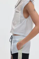 Camisa cropped sin mangas de algodón, con diseño colorblock en tonos blanco y celeste, cuello camisero con ribete negro, cierre frontal de botones y bolsillos en el pecho.