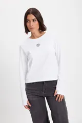 Remera de manga larga ajustada al cuerpo, confeccionada en tejido acanalado color marrón. Presenta cuello redondo, detalle de ojal para el pulgar en los puños y un pequeño bordado decorativo en el centro del pecho.