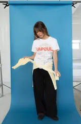 Remera de algodón blanca, con cuello redondo y mangas cortas. Presenta un estampado frontal con la palabra "KAPSUL" en rojo sobre un diseño floral en tono beige.