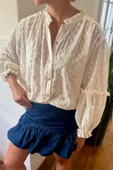 Camisa de corte holgado en color blanco, confeccionada en tela bordada con motivos florales. Presenta cuello mao con volados, cierre frontal con botones de nácar y mangas largas abullonadas con puños elásticos y detalles de volados.