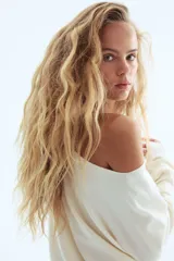 Buzo corto de punto fino en color crema, con diseño de un solo hombro y escote asimétrico. Presenta mangas largas y puños acanalados.