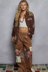 Chaqueta de algodón color marrón con diseño estilo patchwork, que incorpora parches con estampado animal print de leopardo en hombros y bolsillo frontal. Presenta detalles de tachas metálicas, bordes deshilachados, cierre frontal con botones metálicos y un acabado con efecto de pintura desgastada en las mangas.