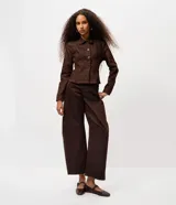 Pantalón de sarga color marrón con corte barrel, cintura media, cierre frontal con botón y cremallera, bolsillos funcionales y recortes estructurales en las piernas.