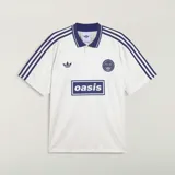 Camiseta Adidas Originals x Oasis de manga corta, color blanco con detalles en azul marino. Presenta cuello polo, las tres tiras características de la marca en los hombros, el logo del trifolio y un estampado frontal con la inscripción "oasis".