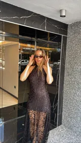 Vestido largo de encaje negro con cuello halter y espalda descubierta. Tiene un corte a la altura de la rodilla y un ruedo largo transparente.