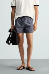 Bañador largo de hombre color rosa pálido con textura seersucker. Presenta cintura elástica ajustable con cordón blanco, bolsillos laterales y un bolsillo trasero de plastrón.