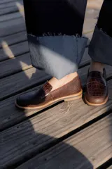 Mocasines negros de cuero con textura tipo cocodrilo en la parte superior y acabado brillante. Presentan una banda frontal clásica y una suela plana con detalle de borde en color natural.