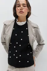 Chaleco negro tejido en lambswool con textura de trenzas y pequeñas flores blancas bordadas a mano dispersas por toda la prenda. Presenta cuello redondo y terminaciones acanaladas.