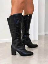 Bota de caña alta confeccionada en cuero negro, con punta almendrada y taco cuadrado de 7 cm. Presenta un diseño de caña amplia con cierre lateral interno y tirador metálico.