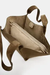 Cartera shopper de hombro confeccionada en gamuja color marrón, con diseño estructurado de laterales abiertos y doble asa superior. Cuenta con cierre de botón imantado e incluye un bolsillo interior extraíble con cremallera.