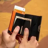 Billetera tarjetero de cuero sintético color negro con costuras en contraste color naranja. Cuenta con cierre lateral mediante broche a presión y diseño compacto.