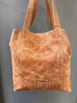 Bolso tipo tote de cuero vacuno en color marrón con acabado desgastado. Presenta costuras a la vista en hilo claro, doble asa de hombro y etiqueta de cuero con logo grabado en la parte inferior frontal.
