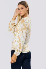 Blusa de manga larga con estampado floral en tonos blanco y mostaza. Presenta escote en V con botones frontales, frunces en los hombros y puños abotonados con detalle de pin metálico con logo.