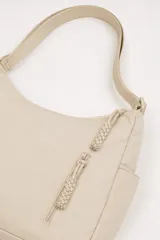 Cartera tipo hobo de nylon color beige, con correa ajustable para el hombro, cierre principal superior y dos bolsillos frontales verticales con cierre y tiradores trenzados. Cuenta además con bolsillos laterales abiertos.