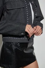Campera de denim color negro con detalles de tachas metálicas en cuello, solapas, solapa de cierre y bolsillos frontales. Posee cierre frontal con botones metálicos y bolsillos con solapa en el pecho.