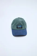Gorra tipo dad hat de pana combinada en bloques de color verde y azul, con visera curvada y un parche bordado en el frente con diseño de montañas y la inscripción "UNIFORM".