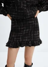 Minifalda negra de tweed con diseño a cuadros blancos, bajo asimétrico con volantes y cierre lateral con cremallera invisible.