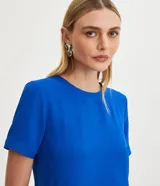 Blusa blanca de poliéster con cuello redondo, mangas cortas, cierre trasero en forma de gota, pequeñas aberturas laterales y acabados en pespuntes en las mangas y el cuello.