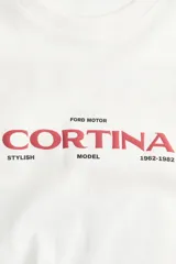 Remera de corte oversized en color blanco, con cuello redondo y mangas cortas de hombros caídos. Presenta un estampado gráfico central con la inscripción 'CORTINA' en color rojo y detalles en negro.