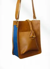 Bolso tipo tote o matero de cuero color camel con laterales en cuero texturado azul eléctrico. Posee un bolsillo frontal exterior y un accesorio colgante de cuero trenzado con un botón de ajuste. Incluye correa de hombro desmontable.