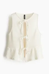 Top corto sin mangas de punto color blanco roto, con escote redondo y abertura frontal que se cierra mediante tres lazos anudados. Presenta un detalle de peplum con ligero vuelo en el bajo.