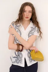 Camisa blanca de manga corta con cuello solapa, cierre frontal de botones y diseño bordado en hilo azul marino con motivos florales y vegetales.